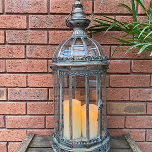 May include: A tall, ornate metal lantern with a glass cage and three white pillar candles inside. The lantern has a distressed, antique finish and intricate details.