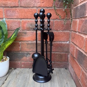 May include: A black fireplace tool set with a stand. The set includes a poker, shovel, brush, and tongs. The tools have decorative handles and are arranged in front of a red brick wall.