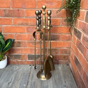 May include: A vintage brass fireplace tool set. The set includes a poker, shovel, and tongs, all with decorative handles. The tools are displayed on a stand with a round base. The background is a brick wall.
