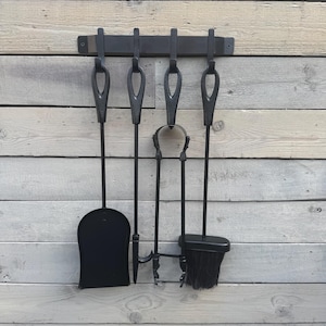 May include: A black fireplace tool set hanging on a dark metal bar. The set includes a shovel, poker, tongs, and a brush. The tools have loop handles and are mounted on a wooden wall.
