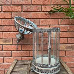 May include: A silver metal lantern with a glass top and intricate detailing. The lantern is open and the glass top is resting on the side. The lantern is sitting on a wooden surface.