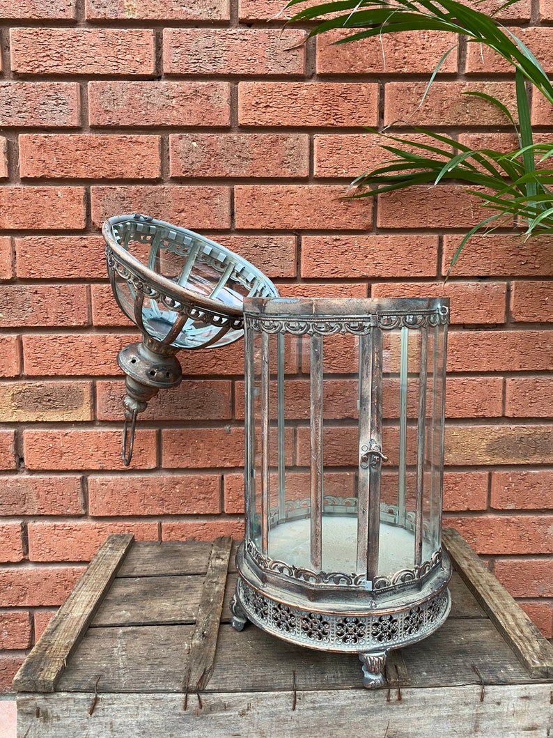May include: A silver metal lantern with a glass top and intricate detailing. The lantern is open and the glass top is resting on the side. The lantern is sitting on a wooden surface.