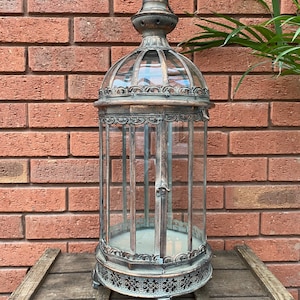 May include: A tall, ornate metal lantern with a glass cage and a decorative top. The lantern has a distressed, antique finish and sits on a wooden surface.