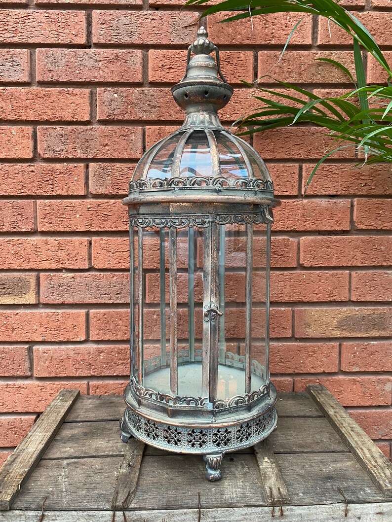 May include: A tall, ornate metal lantern with a glass cage and a decorative top. The lantern has a distressed, antique finish and sits on a wooden surface.