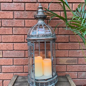 May include: A tall, silver metal lantern with a glass cage and ornate details. The lantern has two white pillar candles inside, creating a warm glow.