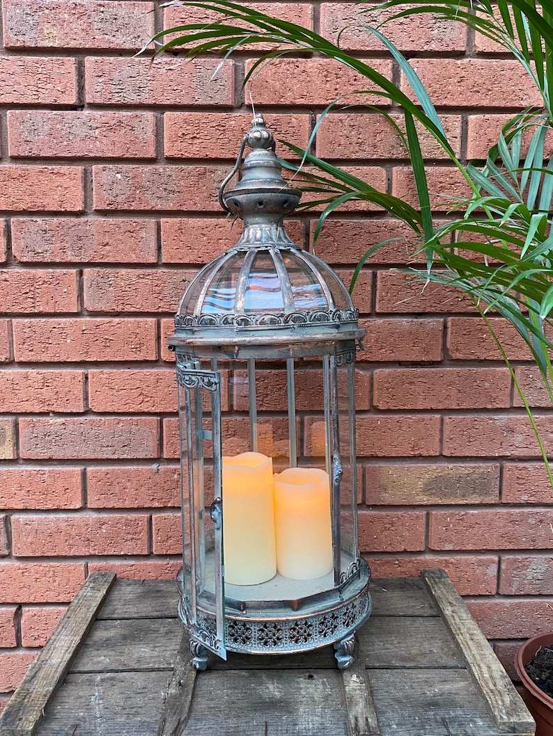 May include: A tall, silver metal lantern with a glass cage and ornate details. The lantern has two white pillar candles inside, creating a warm glow.