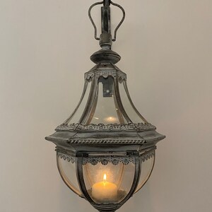 K&ouml;nnte beinhalten: Eine wei&szlig;e Metall-Kerzenlaterne mit Glasgeh&auml;use und dekorativem Design. Die Laterne h&auml;ngt an einem Haken an einer wei&szlig;en Wand und hat eine brennende Kerze im Inneren.