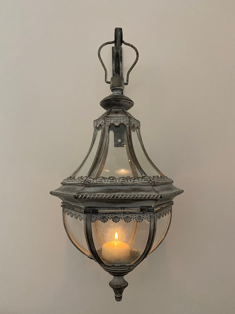 K&ouml;nnte beinhalten: Eine wei&szlig;e Metall-Kerzenlaterne mit Glasgeh&auml;use und dekorativem Design. Die Laterne h&auml;ngt an einem Haken an einer wei&szlig;en Wand und hat eine brennende Kerze im Inneren.