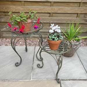 May include: Two metal plant stands with ornate designs, each holding a potted plant. The stand on the left has a fuchsia plant with pink and purple flowers. The stand on the right has a white cyclamen plant. Both stands are on a grey stone patio.