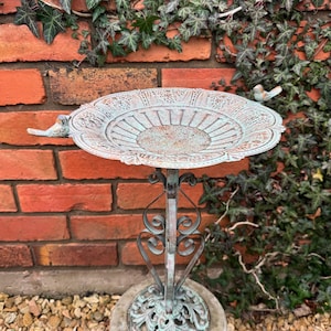 May include: A decorative metal bird bath with a distressed green finish. The bath has a scalloped edge and a raised centre. Two small metal birds are perched on the rim of the bath. The bath is mounted on a metal stand with a scrolled design.