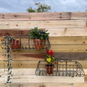 May include: Two metal wall planters with a rustic finish. The top planter has a trailing plant with green leaves and a potted plant with purple flowers. The bottom planter has a potted plant with red and yellow flowers.