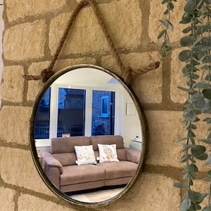 May include: A round, gold-toned mirror with a rope hanger. The mirror is reflecting a brown sofa with two cushions.