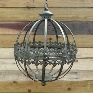 May include: A large, round, metal candle lantern with a decorative, ornate design. The lantern is hanging from a chain and has a distressed, aged finish.