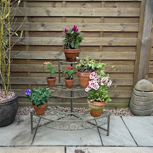 May include: A three-tiered metal plant stand in an antique style, displaying terracotta pots filled with diverse flowering plants. The stand has a curved design with decorative scrollwork. The plants include purple, red, and white flowers, and green foliage. A wooden fence forms the backdrop.