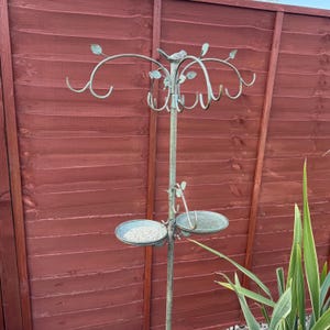 May include: A metal bird feeder with a decorative tree design and two bowls. The feeder is mounted on a pole and has multiple hooks for hanging additional feeders.