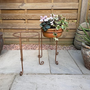 May include: Two rusty metal plant stands with a round top and three legs. The stand on the right has a terracotta pot with pink and white flowers and green leaves.