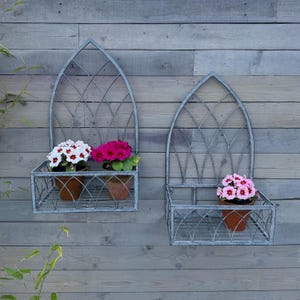 May include: Two grey metal wall planters with arched tops and a diamond pattern. Each planter has a shelf and is holding a potted plant with pink and white flowers.