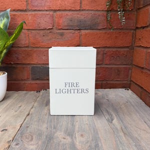 May include: A white rectangular box with the text "FIRE LIGHTERS" in grey. The box is on a wooden surface, set against a red brick wall. A green plant is visible on the left. The box is likely for storing fire lighters.