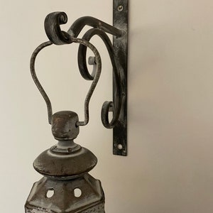 K&ouml;nnte beinhalten: Eine wei&szlig;e und braune, abgenutzte Metall-Laterne mit einer Glasscheibe und einer dekorativen Metallhalterung. Die Halterung ist an einer Wand befestigt.