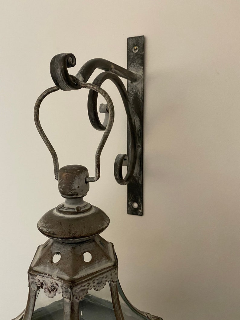 K&ouml;nnte beinhalten: Eine wei&szlig;e und braune, abgenutzte Metall-Laterne mit einer Glasscheibe und einer dekorativen Metallhalterung. Die Halterung ist an einer Wand befestigt.