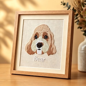 May include: Embroidered portrait of a dog named Bear, framed in a light wood frame. The dog has brown and cream-colored fur, with a detailed stitched design. The artwork is displayed on a beige fabric background.