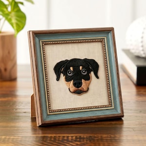 Könnte beinhalten: Gesticktes Rottweiler-Hundeporträt in einem Holzrahmen mit blau-goldener Umrandung. Das Gesicht des Hundes ist schwarz, hellbraun und braun, vor einem neutralen Hintergrund. Der Rahmen steht auf einer Holzoberfläche.