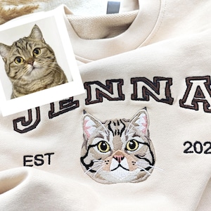 Puede incluir: Sudadera color crema con el nombre "JENNA" bordado en letras marrones. Debajo del nombre, una cara de gato bordada y el año "2021". Una pequeña foto de un gato está en la esquina superior izquierda.