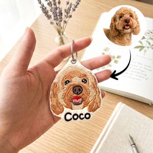 May include: Embroidered keychain featuring a brown dog with the name "Coco" in black lettering. The keychain has a silver ring and a detailed image of the dog's face. A photo of the dog is in the background.