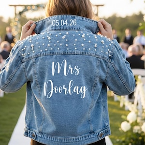 May include: Light blue denim jacket with pearl embellishments on the shoulders. The back of the jacket features the text "Mrs Doorlag" in white script, with the date "05.04.26" above the collar. A wedding-themed garment.