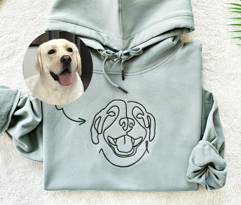 May include: A light green hooded sweatshirt with a black embroidered outline of a dog's face. The dog's face is smiling with its tongue sticking out. A photo of a yellow Labrador Retriever is in the background.