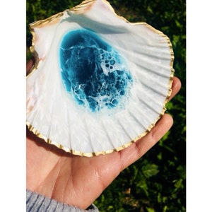 May include: A white seashell with a gold rim, filled with blue resin with white swirls. The seashell is being held in a hand.