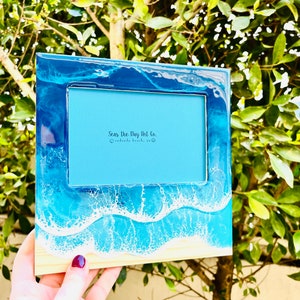 May include: A rectangular photo frame with a beach-themed design. The frame features a blue and white resin wave pattern, with a light blue interior and the text "Seas the Day Art Co." The frame is made of wood and resin.