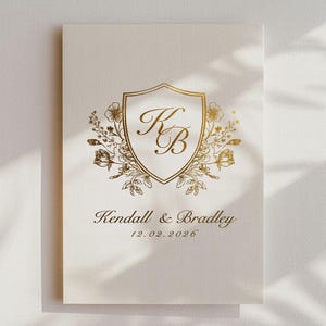 May include: Cream invitation card with gold foil detailing. It features a gold shield emblem with the initials "KB" and floral accents. The names "Kendall & Bradley" and the date "12.02.2026" are printed below the emblem.