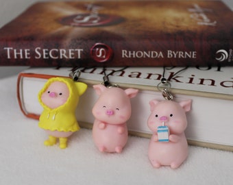 May include: Three pig charm keychains. One pig is wearing a yellow raincoat with a hood. One pig is pink and has a happy expression. One pig is pink and is holding a small blue and white carton.
