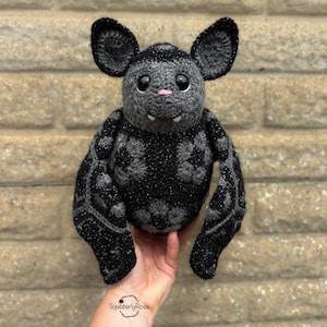 May include: A handmade crochet bat in shades of gray and black. The bat has large ears, a pink nose, and small fangs. The wings and body feature a detailed pattern. The bat is held in a hand, with the maker's name, SquibberlyRose, visible at the bottom.