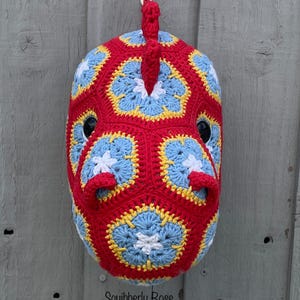 May include: A handmade, crocheted horse mask in red, blue, yellow, and white. The mask features a detailed floral pattern and black eye openings. The text "Squibberly Rose" is visible on the bottom of the mask.
