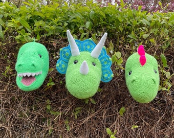May include: Three crocheted dinosaur heads, a green T-Rex with teeth, a green Triceratops with blue and white accents, and a green Stegosaurus with a pink spike, are displayed against a green bush background.