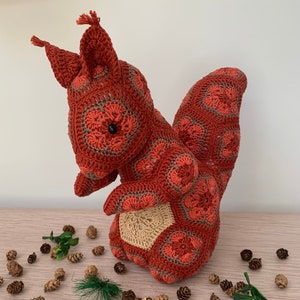 May include: A crocheted red squirrel toy with a brown tail and a brown patch on its back. The squirrel is surrounded by small pine cones and green sprigs.