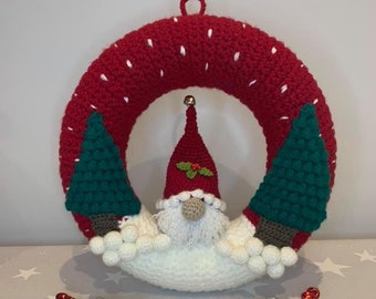 Op de afbeelding: Een gehaakte kerstkrans met een kabouter in het midden. De krans is rood met witte stippen en heeft twee groene bomen aan weerszijden van de kabouter. De kabouter draagt ​​een rode muts met een witte pompon en een witte baard.