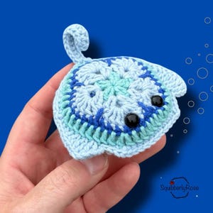 Könnte beinhalten: Eine handgehäkelte Rochenfigur in Blau- und Türkistönen mit schwarzen Perlenaugen. Das Seeungeheuer hat einen eingerollten Schwanz und wird vor einem blauen Hintergrund gehalten. Das Bild enthält den Text "SquibblyRose".