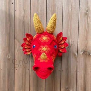 Puede incluir: Una cabeza de dragón de crochet roja y amarilla colgada en la pared. El dragón tiene ojos azules y un diseño texturizado y escamoso.