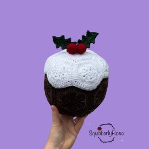 May include: A crocheted Christmas pudding ornament with white icing, brown cake, and green holly with red berries. The ornament is being held in a hand.