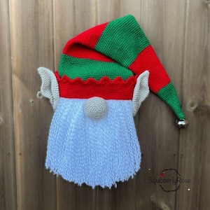May include: A crocheted elf hat with a white beard, red and green stripes, and a bell on the end. The hat is hanging on a wooden fence.
