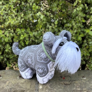 Könnte beinhalten: Handgehäkelter Schnauzer-Hund aus grauem und weißem Garn. Der Hund hat einen weißen Bart, schwarze Augen und ein grünes Halsband. Der Häkelhund sitzt auf einer Steinmauer vor grünem Laub.