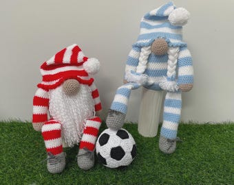 May include: Two crocheted gnomes with long legs. One gnome wears a red and white striped hat and has a white beard. The other gnome wears a blue and white striped hat with white braided hair. A black and white crocheted soccer ball sits between them.