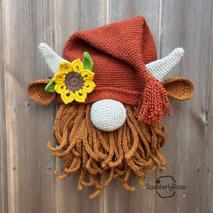 May include: A crocheted gnome wall hanging featuring a rust-colored pointed hat with a tassel, two light-colored horns, brown ears, a yellow sunflower with a green leaf, a round light-colored nose, and a long, curly brown yarn beard.