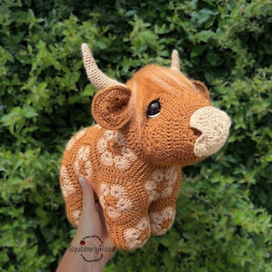 May include: A crocheted Highland cow figurine in shades of brown and tan. The cow has detailed features, including horns, ears, and a fluffy head. The body is decorated with a floral pattern. The figurine is held in front of a green leafy background.