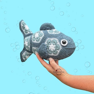 May include: A stuffed fish toy crafted from crocheted yarn in shades of slate blue, sage green, and white. The body is constructed from multiple hexagonal motifs featuring a central white flower pattern. It has a single dark eye with a white border.