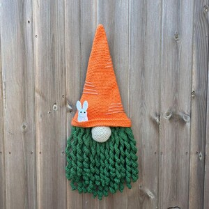 May include: A handmade crochet gnome with an orange carrot-shaped hat and a green beard. The hat has white stripes and a small white bunny applique. The gnome's nose is a light beige sphere. Perfect for Easter decor.