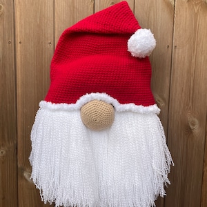 May include: A festive Christmas gnome decoration. It has a red crocheted hat with a white pom-pom, a beige crocheted nose, and a long, white, fluffy beard. This gnome is designed for hanging on a wall or door, perfect for the holidays.